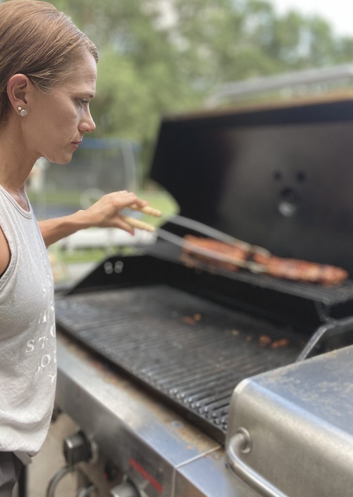 woman grilling on a gas grill using tongs and in a gray tank top